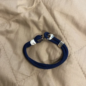 Marinblått rep-armband med silverfärgade detaljer - Säljer ett stilrent armband i marinblått rep med silverfärgade metalldetaljer och vit lindning. Armbandet har en unik krokformad. Perfekt accessoar för en nautisk look.