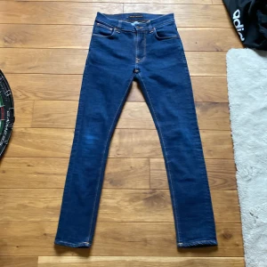 Mörkblå jeans från Nudie Jeans - Säljer ett par mörkblå jeans från Nudie Jeans med klassiska orange sömmar och broderade bakfickor. Modellen har normal passform och raka ben. Perfekta för dig som gillar stilrena och tidlösa jeans. Model Lean Dean och har jeansen är aldrig använda