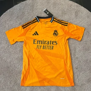 Real Madrid bortatröja 2023/24 från Adidas - Säljer en gul-orange Real Madrid bortatröja från Adidas med svarta detaljer och klubbmärke på bröstet. Tröjan har korta ärmar, rund hals och är tillverkad i lätt funktionsmaterial. Perfekt för fotbollsträning eller att bära som supporter.