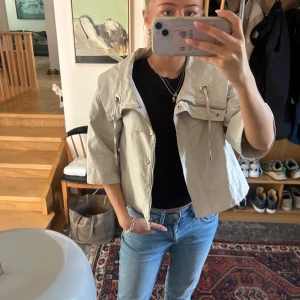 Beige croppad jacka med dragkedja - Säljer en beige croppad jacka med dragkedja och stora fickor framtill. Jackan har trekvartsärm och justerbar dragsko vid kragen. Perfekt för lager-på-lager och snygg till jeans.