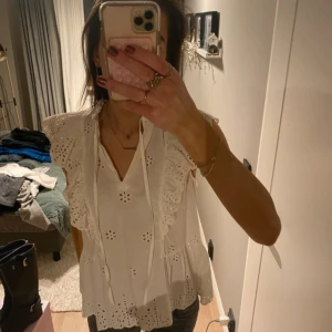 Vit blus med spetsdetaljer och volang - Säljer en söt vit blus med broderade blommor, spetsdetaljer och volang vid axlarna. Blusen har v-ringning med knytband och är ärmlös. Perfekt för vår och sommar!