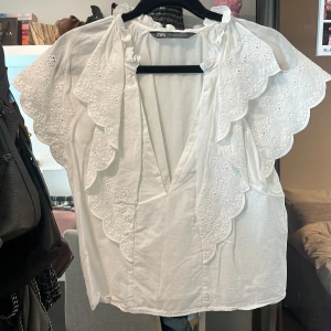 Vit blus med volang från Zara - Säljer en söt vit blus från Zara med broderade volangdetaljer längs v-ringningen och ärmarna. Blusen har korta, vida ärmar och är tillverkad i bomull. Perfekt för vår och sommar!