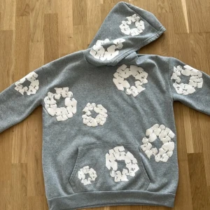 Grå hoodie med vita blommor från Denim Tears - Säljer en grå hoodie från Denim Tears med stora vita blommönster över hela tröjan. Hoodien har magficka och ribbade muddar vid ärmslut och nederkant. Perfekt för dig som gillar streetwear och unika prints. Säljer den för att jag tyckte inte den passade mig bra har haft en en gång och det är på bilder priset kan diskuteras 
