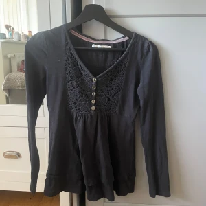 Svart långärmad topp med spets och knappar - Säljer en svart långärmad topp med v-ringning, spetsdetaljer och dekorativa knappar framtill. Toppen har en feminin och lite lös passform, perfekt att styla till olika outfits.