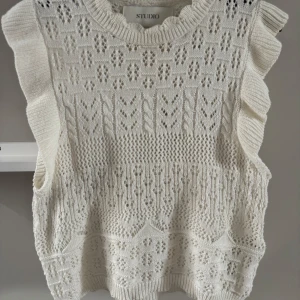 Vit stickad ärmlös topp med volang - Supersöt vit stickad topp från Studio med volangdetaljer längs axlarna och ett fint hålmönster. Perfekt att styla med jeanskjol eller byxor för en fräsch look. Rund halsringning och mjukt material.