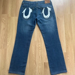True Religion jeans med vita detaljer - Säljer ett par blå jeans från True Religion med snygga vita sömmar och stora loggor på bakfickorna. Jeansen har normal passform och är bootcut, tillverkade i slitstark denim. Midjemått är W28. (USA strl). Det är ungefär 36-38 i EU strl.