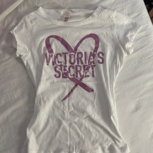 Vit t-shirt från Victoria's Secret med glittertryck - Vit croppad t-shirt från Victoria's Secret med glittrigt rosa tryck och hjärtmotiv på bröstet. T-shirten har korta ärmar och är tillverkad i mjuk bomull. Perfekt för en avslappnad och trendig look.