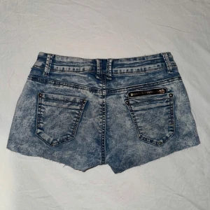 Blå lågmidjade vintage jeansshorts med dragkedjedetalj - Säljer ett par ljusblå jeansshorts med råa kanter och snygg dragkedjedetalj vid ena fickan. Shortsen har klassiska fem fickor och knappstängning framtill. Perfekta för sommaren och ger en cool, avslappnad look. Midjemått: 32cm (strechigt material), grenmåttet 18cm! 🫶🏽💓