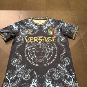 Fotbollströja - Snygg svart fotbollströja med guldiga detaljer och Italiens landslagsemblem. Tröjan har korta ärmar, grekiskt mönster runt kragen och ärmsluten samt stora grafiska mönster i ljusblått. Perfekt för dig som gillar unik stil och fotboll.