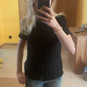 Svart kortärmad topp med struktur - Säljer en svart topp med korta ärmar och unik strukturerad yta. Toppen har rund halsringning och är tillverkad i ett syntetmaterial som ger en cool textur. Perfekt att styla med jeans eller kjol.❤️