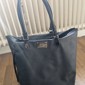Svart shoppingväska från Hunkemöller - Stor svart shoppingväska från Hunkemöller med blanka handtag och guldfärgad logga framtill. Väskan har en enkel design och en liten rem med guldfärgad detalj. Perfekt för att bära med sig allt man behöver under dagen.