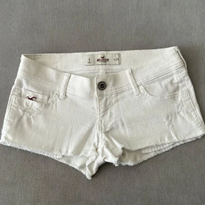 Vita jeansshorts från Hollister - Säljer ett par vita jeansshorts från Hollister med fransig kant och klassisk femficksdesign. Shortsen har låg midja, knapp och dragkedja framtill samt broderad logga på fickan. Perfekta för varma dagar!