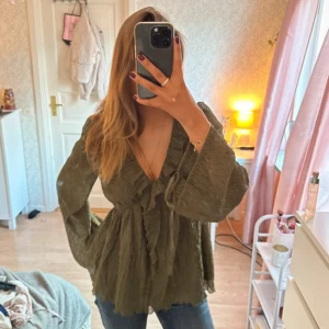 Grön blus med volang och knyt - Säljer en olivgrön blus med v-ringning, volangdetaljer och knyt framtill. Blusen har långa, vida ärmar. Från NA-KD med lappen kvar. Säljer då jag ska flytta och måste rensa garderoben!💕