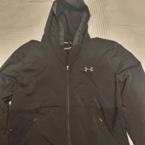 Svart jacka med huva från Under Armour - Svart träningsjacka från Under Armour med huva och dragkedja framtill. Jackan har två sidofickor med dragkedja och diskret logga på bröstet. 