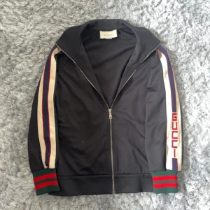 Svart trackjacka från Gucci med ränder - Svart trackjacka från Gucci med dragkedja framtill. Ärmarna har vita, röda och blå ränder samt gröna och röda muddar. På ena ärmen står det 'GUCCI' i rött. Klassisk sportig look med hög krage.