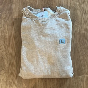 Beige sweatshirt från Les Deux - Säljer en beige sweatshirt från Les Deux med broderad blå logga på bröstet. Tröjan har rund halsringning och är tillverkad i mjukt material med ribbade muddar. Perfekt för en avslappnad stil.