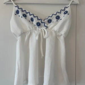 Vit broderad linnetopp med blå detaljer - Säljer en söt vit linnetopp med justerbara axelband och blå broderade blommor längs v-ringningen. Toppen har knytning under bysten och är lätt utsvängd nertill. Den är perfekt till sommaren och är i nytt skick. Varan är från SHEIN och kostar originellt 109. Tveka inte vid frågor!💕
