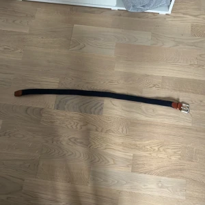 Flätat bälte  - Säljer ett mörkblått flätat bälte från Jack & Jones med bruna läderdetaljer och silverspänne. Bältet har en stilren design och passar perfekt till jeans eller chinos. Längden är märkt 90.