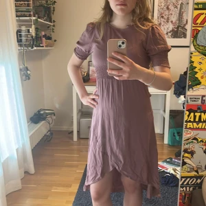 Ljuslila kortklänning med puffärm - Säljer en söt ljuslila klänning med kort ärm och smockdetaljer vid midjan och ärmsluten. Klänningen har en mjuk, flowy passform och lätt v-ringning. Perfekt för sommarens alla tillfällen! använd 1 gång (då jag inte hittar tillfälle till att använda) så ser helt ny ut och köpte för 500 kr