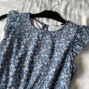 Blommig blå klänning med volangärmar - Supersöt blå klänning med småblommigt mönster i rosa och vitt. Klänningen har volangärmar, rund halsringning och smock i midjan för en bekväm passform. Perfekt för sommarens alla tillfällen!