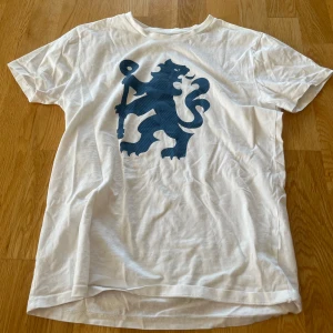 Vit t-shirt med blått lejontryck (Chelsea FC) - Vit t-shirt med ett stort blått lejontryck på framsidan. Klassisk rund hals och korta ärmar. Perfekt för en avslappnad stil och enkel att matcha med jeans eller shorts.