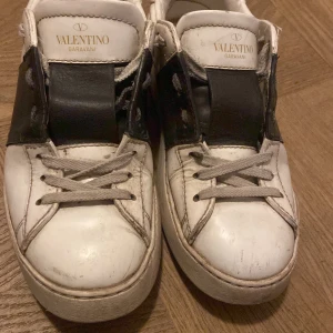 Valentino Garavani sneakers med svart detalj - Säljer ett par vita Valentino Garavani sneakers med bred svart rem över vristen och nitar på hälen. Skorna har rund tå, platt sula och klassisk snörning. Perfekta för dig som gillar exklusiva och stilrena sneakers.