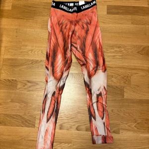 Muskelimmitation  tights från Labellamafia - Säljer ett par unika tights från Labellamafia med röd-orange och vit muskelinspirerat mönster. Byxorna har svart resår i midjan med logga och är tillverkade i stretchigt material som sitter tight.