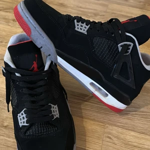 Svarta Nike Air Jordans med röda och grå detaljer - Säljer ett par klassiska Nike Air Jordans i svart mocka med röda och grå detaljer. Skorna har snörning, meshpaneler och en platt sula med synlig luftdämpning. Perfekta för dig som gillar streetwear och sneakers med ikonisk design.