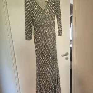 Glittrig långklänning från Mac Duggal - Säljer en superglittrig långklänning från Mac Duggal med v-ringning och långa ärmar. Klänningen är täckt av silverfärgade paljetter i ett rutigt mönster och har en markerad midja. Perfekt för dig som vill sticka ut på festen!