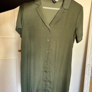 Grön skjortklänning med knappar - Säljer en enkel och stilren grön skjortklänning med korta ärmar och knappar framtill. Klänningen har klassisk krage och är tillverkad i ett mjukt, lätt material som passar perfekt till varmare dagar.