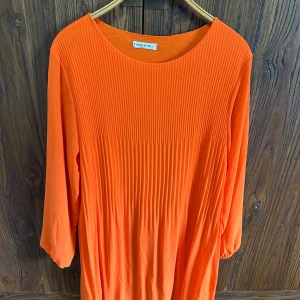 Plisserad orange klänning/lång bluse med trekvartsärm - Säljer en snygg orange klänning med plisserad struktur och rund halsringning. Klänningen har trekvartsärmar och ett luftigt fall, perfekt för dig som gillar färg och stil. Märke: House of Lola.