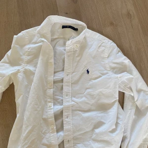 Vit skjorta från Ralph Lauren - Klassisk vit skjorta från Ralph Lauren med blå broderad logga på bröstet. Skjortan har knappar framtill och krage, perfekt för en stilren look. Använd 2 gånger, priset är inte hugget i sten 