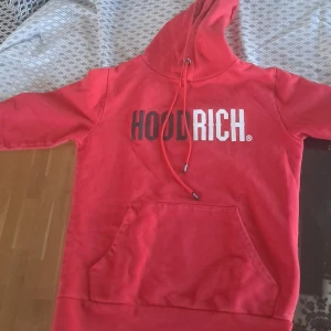 Röd hoodie från Hoodrich - Säljer en röd hoodie från Hoodrich med vit och svart logga på bröstet. Tröjan har huva med dragsko och en stor magficka framtill. Perfekt för dig som gillar streetwear och vill sticka ut med en färgstark look. Den har även en kedja vid armen