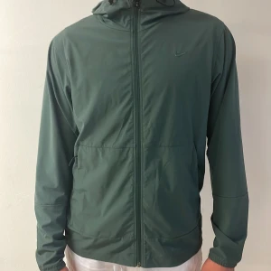 Grön vindjacka från Nike - Snygg grön vindjacka från Nike med huva och dragkedja framtill. Jackan har två sidofickor och diskret Nike-logga på bröstet. Perfekt för blåsiga dagar och enkel att matcha med olika outfits.