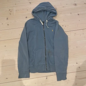 Blå hoodie från Polo Ralph Lauren - Säljer en blå hoodie från Polo Ralph Lauren med dragkedja och huva. Tröjan har en gul broderad logga på bröstet, fickor framtill och ribbade muddar vid ärmslut och nederkant. Perfekt för en avslappnad stil. Skick 8/10. Storlek XL men passar även L, skriv om ni undrar något!