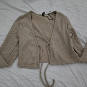 Beige ribbad omlottkofta från H&M Divided - Säljer en beige ribbad omlottkofta från H&M Divided i storlek XS. Koftan har långa ärmar och knytband framtill, perfekt för lager-på-lager. Enkel och stilren design som passar till mycket.