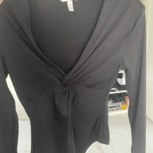 Svart body  - Säljer en svart omlottopp från H&M med lång ärm och snygg v-ringning. Toppen har twistad detalj framtill och är i ett mjukt, stretchigt material. Perfekt till många olika outfits!