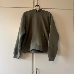 Olivgrön hoodie Hilma XS - Olivgrön hoodie från Hilma i storlek XS. Tröjan har huva, magficka och ribbade muddar vid ärmslut och nederkant. Perfekt för en chill och avslappnad stil. Materialet är mjukt och känns skönt mot huden.