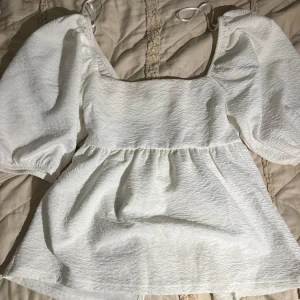 Vit blus med puffärm från Gina Tricot - Supersöt vit blus från Gina Tricot i storlek XS. Blusen har puffiga trekvartsärmar, fyrkantig ringning och knytband i ryggen. Materialet är strukturerat och ger en lyxig känsla. Perfekt för dig som gillar romantisk och trendig stil.