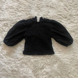 Svart blus med puffärmar från H&M Divided - Supersnygg svart blus från H&M Divided med puffiga ärmar. Toppen har en liten volangkant nertill och hög hals med knapp baktill. Perfekt för dig som gillar trendiga detaljer och en cool siluett!😍