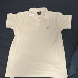 Vit pikétröja från GANT i bomull - Klassisk vit pikétröja från GANT i storlek L. Tröjan har korta ärmar, krage och knäppning framtill samt GANT-logga broderad på bröstet. Tillverkad i mjuk bomull som andas, perfekt för en clean och stilren look.