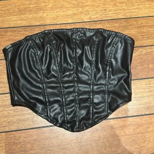 Svart korsett i skinnimitation - Säljer en svart korsett i skinnmaterial med markerade sömmar och dragkedja bak. Toppen är axelbandslös och perfekt för att lyfta vilken outfit som helst. 
