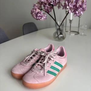 Adidas Gazelle rosa/gröna sneakers - Säljer ett par Adidas Gazelle sneakers i ljusrosa mocka med gröna ränder och detaljer. Skorna har rosa hälparti med grönt Adidas-logo, klassisk gummisula och snörning. Perfekt för dig som vill sticka ut med färg och retrostil.