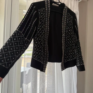Svart cardigan jacka med pärlor Cubus - Svart kavaj jacka från Cubus med långa ärmar och täckt av silvriga pärlor i rutmönster och längs kanterna. Jackan är öppen framtill och har en cool, statement look som verkligen sticker ut. Perfekt för dig som vill ha något extra till outfiten. Alla pärlor är intakta, vissa kan va ”lösare” än andra. Men sitter på sin plats!