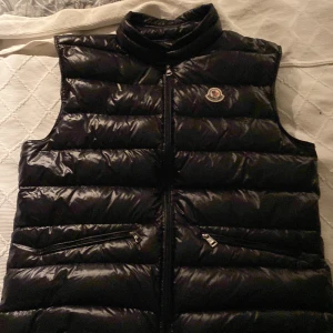 Svart dunväst från Moncler(pris kan diskuteras) - Säljer en svart, glansig dunväst från Moncler Longue Saison. Västen har hög krage, två snedställda fickor med dragkedja och klassisk Moncler-logga på bröstet. Perfekt för lager-på-lager och riktigt snygg till streetwear eller casual outfits.