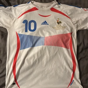 Frankrike Zidane VM 2006   - Säljer en vit Frankrike t shirt från Adidas med blå och röda detaljer. Tröjan har korta ärmar och nummer 10 på framsidan samt namnet 'Zidane' på baksidan. Från final 2006 Italien vs Frankrike. LIMITED EDITION! Haft den bara hängd uppe i rummet. 