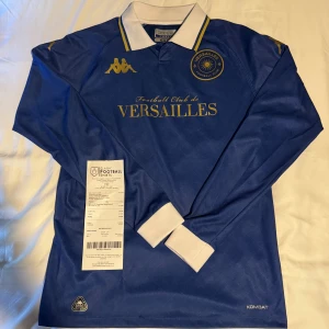 FC Versailles långärmad matchtröja Kappa - Snygg blå långärmad FC Versailles fotbollströja från Kappa med vita muddar och krage, samt guldiga detaljer och klubbmärke på bröstet. Klassisk design med tryckt logga och text, tillverkad i lätt och ventilerande material. Perfekt för dig som gillar stilrena retrovibbar.