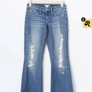 Blå bootcut jeans med slitningar - Snygga blå bootcut jeans med slitna detaljer framtill och coola bakfickor med dekorativa sömmar och knappar. Jeansen har låg midja och klassisk femficksdesign. Perfekta för dig som gillar en avslappnad och trendig look.