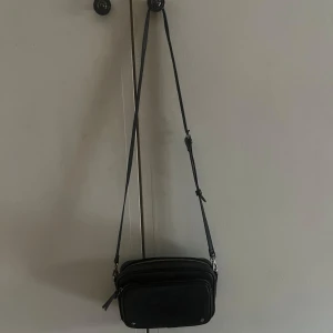 Svart axelväska i skinn med dragkedjor - Snygg svart axelväska i skinn med flera fack och silverfärgade dragkedjor. Väskan har justerbar rem och stilren design som passar till det mesta. Perfekt storlek för mobil, plånbok och andra småsaker!