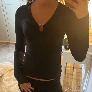 Svart långärmad topp med v-ringning - Säljer en svart långärmad topp med v-ringning och snygg rynkdetalj framtill. Toppen har en liten cut-out vid bysten och sitter tight mot kroppen. Perfekt för dig som gillar en stilren men ändå trendig look.